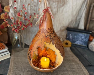 A dried gourd with a carved opening to hold a battery operated candle. Foliage artwork is added to the gourd shell.